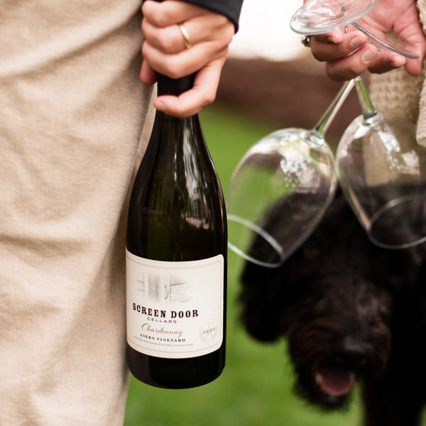 One person holding a bottle of Screen Door Cellars Chardonnay, a dog and a second person carrying two empty wine glasses.