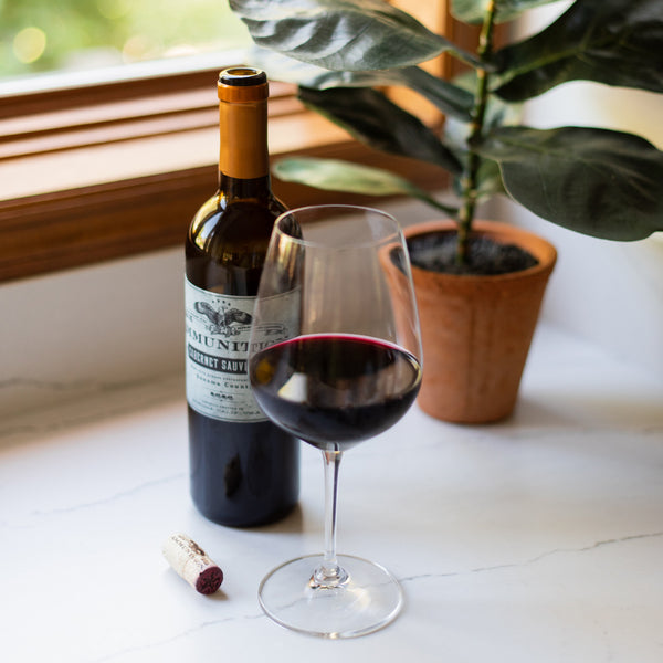 A bottle of Ammunition Cabernet Sauvignon and a partially filled glass of wine on a counter in front of a window and potted plant.